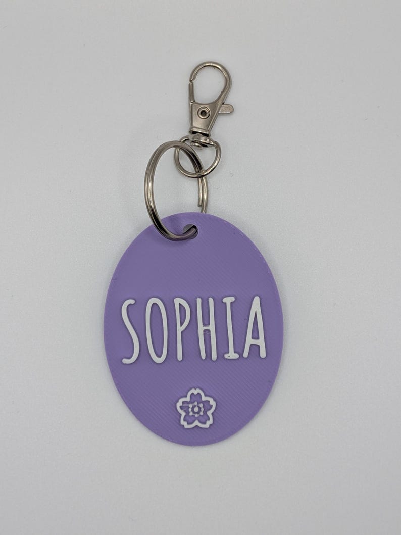 May include: A lavender oval keychain with the name "SOPHIA" in white, accompanied by a small flower graphic. The keychain has a silver-colored clasp and ring for attachment.
