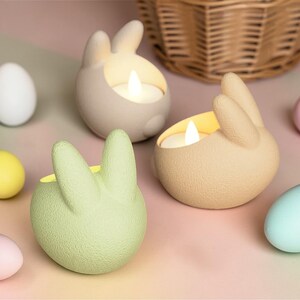 K&ouml;nnte beinhalten: Osterdekoration mit drei hasenf&ouml;rmigen Kerzenhaltern in Beige und Gr&uuml;n, jeweils mit einer brennenden Kerze. Pastellfarbene Eier in Wei&szlig;, Gelb, Rosa und Blau umgeben die Kerzenhalter, mit einem Weidenkorb im Hintergrund.