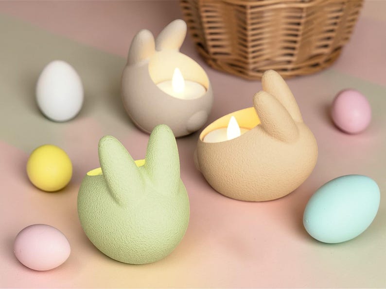 K&ouml;nnte beinhalten: Osterdekoration mit drei hasenf&ouml;rmigen Kerzenhaltern in Beige und Gr&uuml;n, jeweils mit einer brennenden Kerze. Pastellfarbene Eier in Wei&szlig;, Gelb, Rosa und Blau umgeben die Kerzenhalter, mit einem Weidenkorb im Hintergrund.