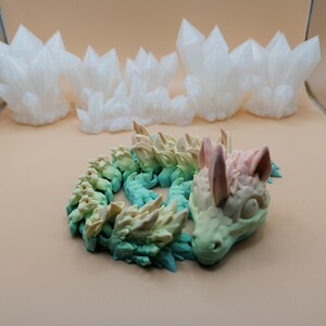 Puede incluir: Una figura de drag&oacute;n en espiral multicolor con un degradado de tonos verdes, azules y amarillos. La cabeza del drag&oacute;n es de color rosa p&aacute;lido y verde. En el fondo hay varias formaciones de cristales blancos.