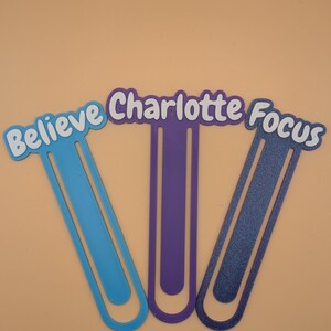 May include: Three plastic bookmarks shaped like large paperclips. The left bookmark is light blue with the word "Believe" in white. The centre bookmark is purple with the name "Charlotte" in white. The right bookmark is dark blue with the word "Focus" in white.