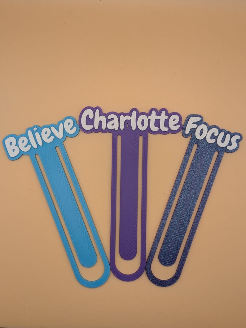 May include: Three plastic bookmarks shaped like large paperclips. The left bookmark is light blue with the word "Believe" in white. The centre bookmark is purple with the name "Charlotte" in white. The right bookmark is dark blue with the word "Focus" in white.