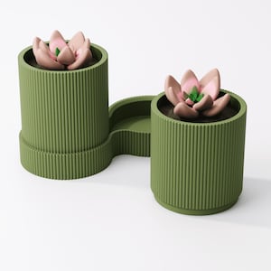 May include: Olive green planter with two cylindrical pots connected by a curved tray. Each pot contains a faux succulent with pink and green petals. The planter has vertical ribbed detailing.