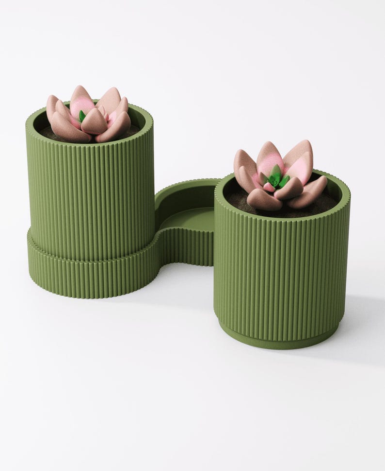 May include: Olive green planter with two cylindrical pots connected by a curved tray. Each pot contains a faux succulent with pink and green petals. The planter has vertical ribbed detailing.