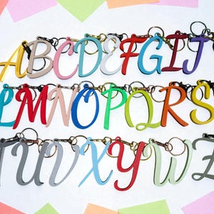 May include: A collection of colorful, cursive letter keychains, each with a silver keyring. The letters are in various colors, including yellow, pink, blue, red, green, and gray. The keychains are arranged in rows, spelling out the alphabet.