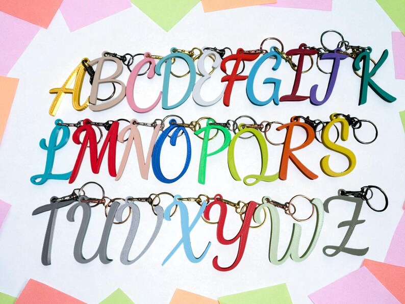 May include: A collection of colorful, cursive letter keychains, each with a silver keyring. The letters are in various colors, including yellow, pink, blue, red, green, and gray. The keychains are arranged in rows, spelling out the alphabet.