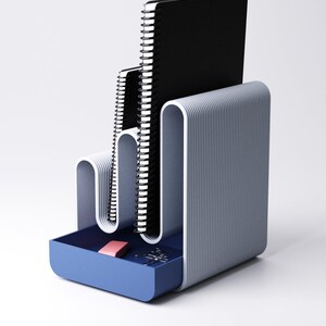 May include: A modern desk organizer in shades of gray and blue. It features a tiered design to hold notebooks and a small drawer containing a pink eraser and paper clips. The organizer has a minimalist, streamlined aesthetic.