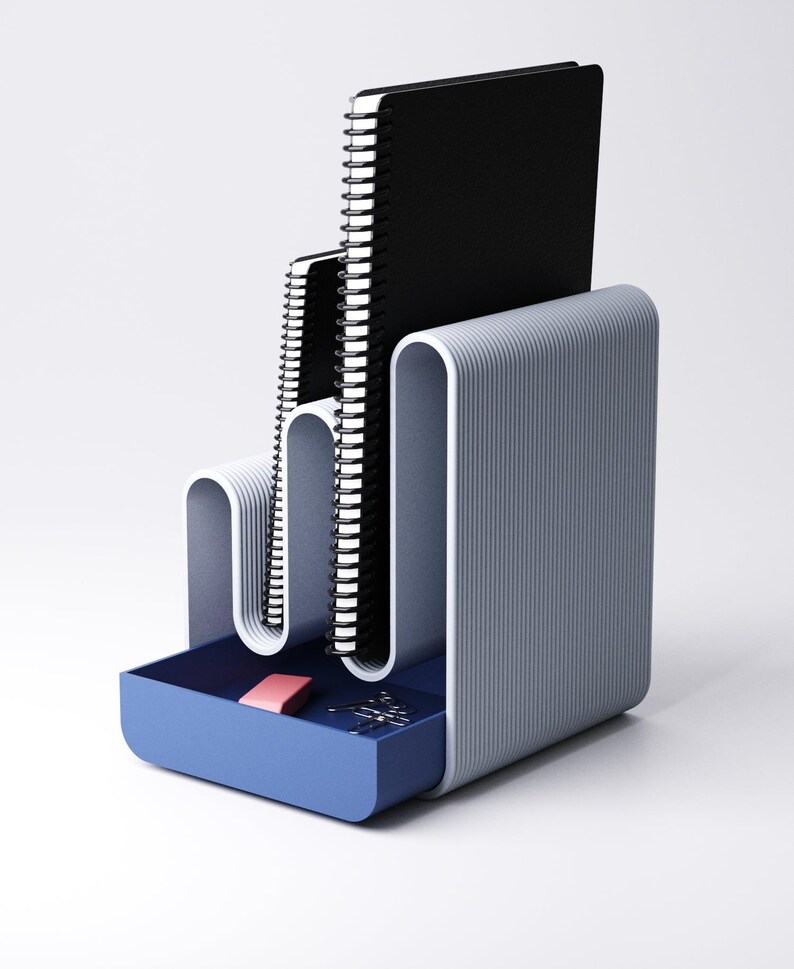 May include: A modern desk organizer in shades of gray and blue. It features a tiered design to hold notebooks and a small drawer containing a pink eraser and paper clips. The organizer has a minimalist, streamlined aesthetic.
