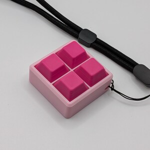 May include: A pink square-shaped fidget toy with four raised pink squares inside a light pink frame. A black lanyard is attached to the toy, with a black slider and a gray end piece. The background is white.