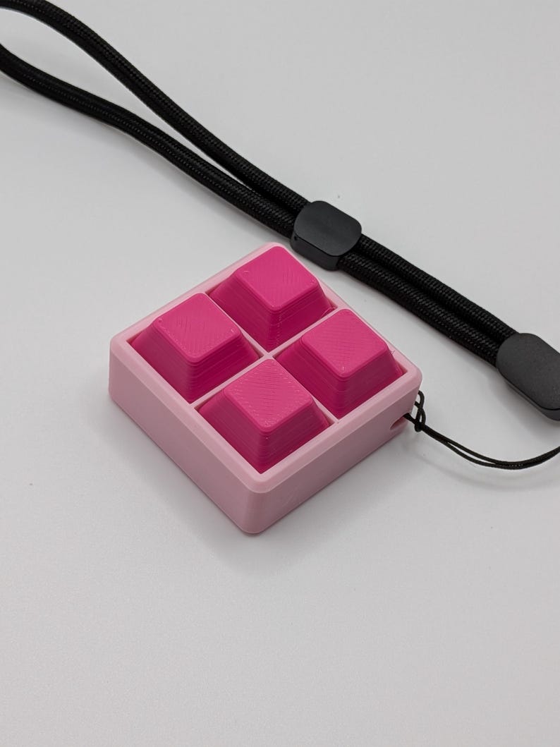 May include: A pink square-shaped fidget toy with four raised pink squares inside a light pink frame. A black lanyard is attached to the toy, with a black slider and a gray end piece. The background is white.
