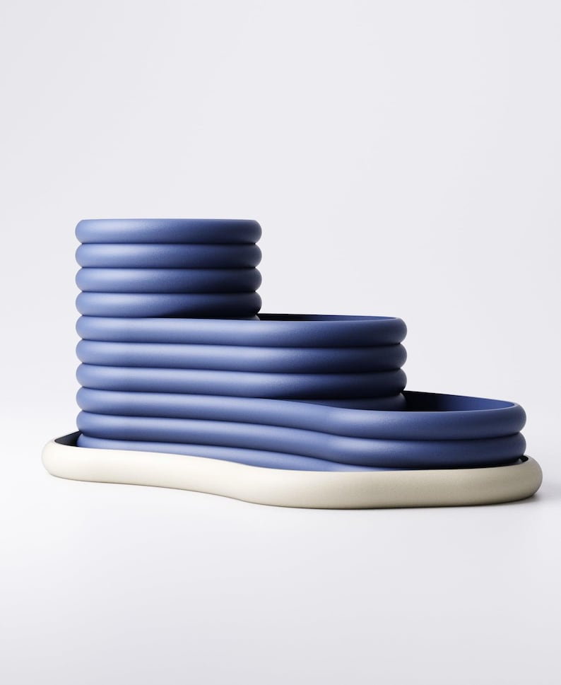 May include: A modern, blue ceramic decorative object with a tiered design. The object features a series of stacked, rounded forms in varying heights, resting on a white base. The overall shape is geometric and sculptural, suitable for home decor.