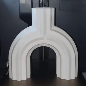 May include: A white, arch-shaped decorative object with a vertical top section and a rounded base. The design features layered, curved lines, creating a modern, geometric aesthetic. The object is likely 3D-printed.