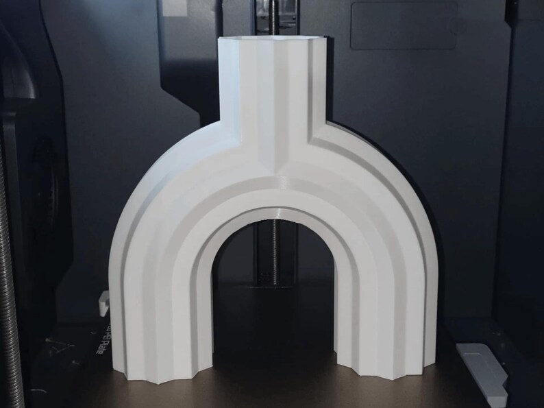 May include: A white, arch-shaped decorative object with a vertical top section and a rounded base. The design features layered, curved lines, creating a modern, geometric aesthetic. The object is likely 3D-printed.