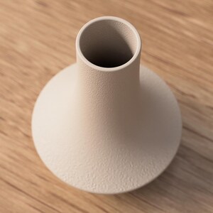 May include: A beige ceramic vase with a textured surface. The vase has a rounded base and a cylindrical neck. The vase is sitting on a wooden surface.