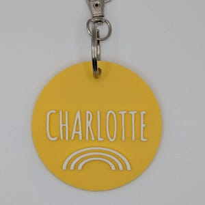 May include: A yellow circular keychain with the name "CHARLOTTE" in white block letters and a white rainbow graphic. The keychain has a silver clasp and a split ring for attaching keys.