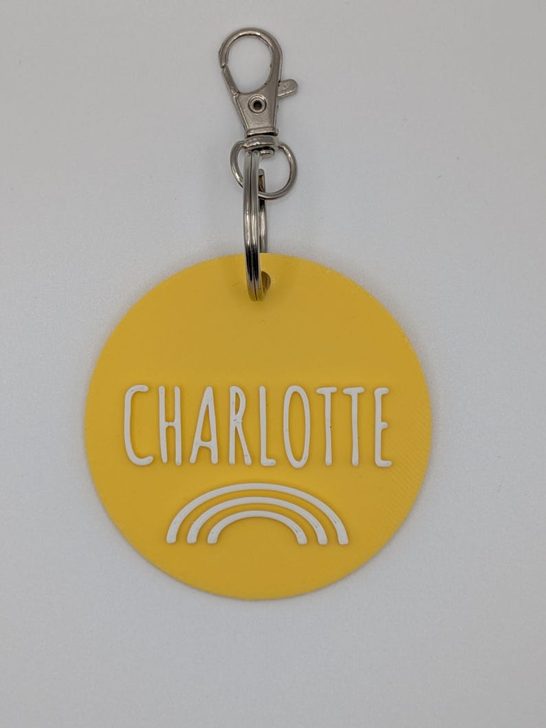 May include: A yellow circular keychain with the name "CHARLOTTE" in white block letters and a white rainbow graphic. The keychain has a silver clasp and a split ring for attaching keys.