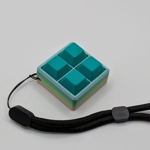 May include: A teal and blue square-shaped fidget toy with four raised squares. The toy has a gradient edge of pink, yellow, and blue. A black wrist strap is attached to the toy.