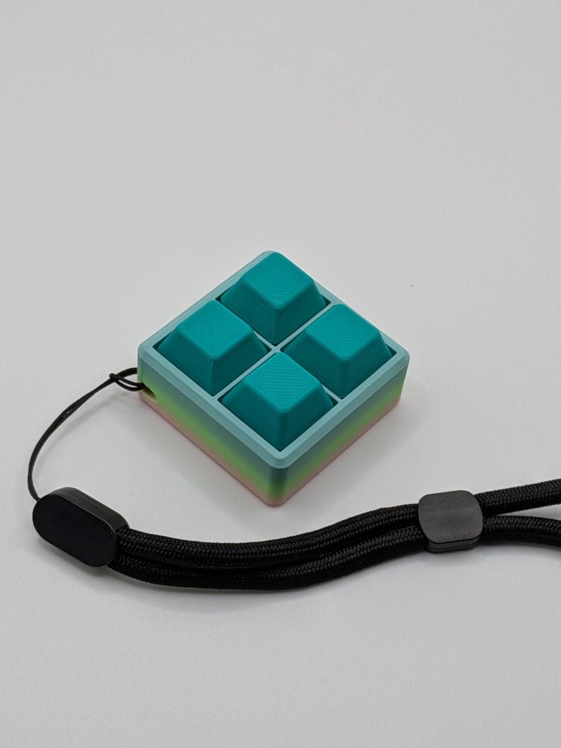 May include: A teal and blue square-shaped fidget toy with four raised squares. The toy has a gradient edge of pink, yellow, and blue. A black wrist strap is attached to the toy.