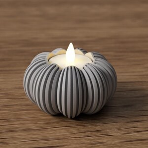 May include: A gray and white striped pumpkin-shaped candle holder with a lit candle. The candle holder is made of ceramic or a similar material and sits on a wooden surface. The candle flame is bright white.