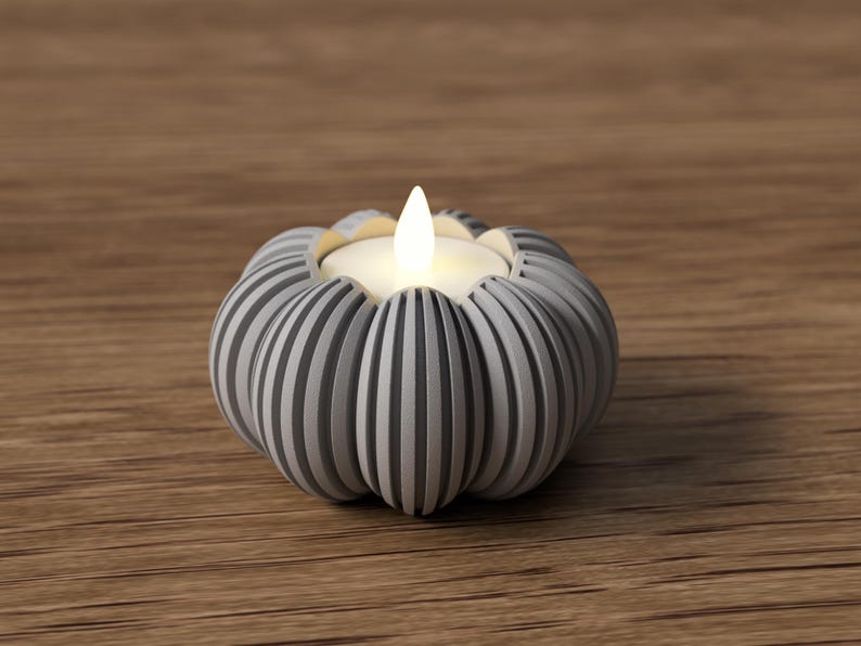May include: A gray and white striped pumpkin-shaped candle holder with a lit candle. The candle holder is made of ceramic or a similar material and sits on a wooden surface. The candle flame is bright white.