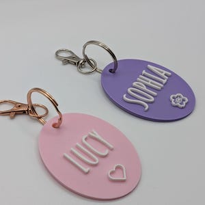 May include: Two personalized keychains. One is pink with the name "LUCY" and a heart, the other is purple with the name "SOPHIA" and a flower. Both have metal key rings and clasps.
