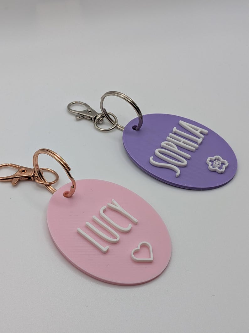 May include: Two personalized keychains. One is pink with the name "LUCY" and a heart, the other is purple with the name "SOPHIA" and a flower. Both have metal key rings and clasps.