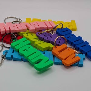 May include: A collection of colorful 3D-printed name keychains in various colors, including pink, yellow, green, blue, and orange. Each keychain features a different name, attached to a silver keyring.