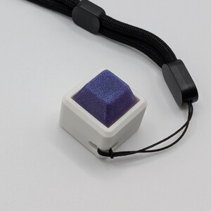 May include: A white square-shaped object with a dark blue, textured top. The object is attached to a black lanyard. The lanyard has a black plastic adjuster and a black cord. The background is white.