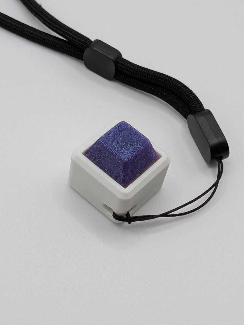 May include: A white square-shaped object with a dark blue, textured top. The object is attached to a black lanyard. The lanyard has a black plastic adjuster and a black cord. The background is white.