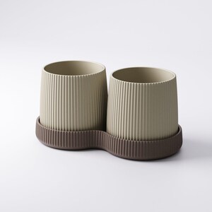 May include: Two beige cylindrical containers with vertical ribbed detailing, set on a brown, wavy-edged base. The containers have a minimalist design, suitable for holding items like pens or small plants. The neutral color palette offers a modern aesthetic.