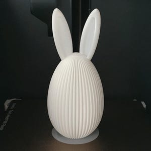 Puede incluir: Objeto decorativo blanco en forma de huevo con detalles verticales acanalados. Dos orejas redondeadas y erguidas se extienden desde la parte superior, dando la apariencia de un conejo. El objeto est&aacute; sobre una base circular, sobre un fondo oscuro.