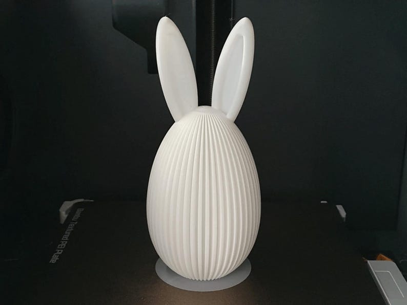 Puede incluir: Objeto decorativo blanco en forma de huevo con detalles verticales acanalados. Dos orejas redondeadas y erguidas se extienden desde la parte superior, dando la apariencia de un conejo. El objeto est&aacute; sobre una base circular, sobre un fondo oscuro.
