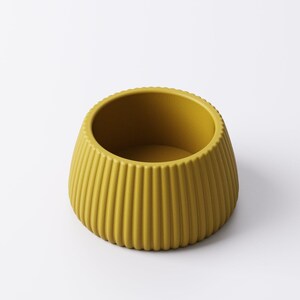May include: Mustard yellow ceramic bowl with vertical ribbed detailing. The bowl has a rounded shape with a slightly flared base and a smooth interior. The rim has a subtle, textured edge. This decorative bowl is suitable for various uses.