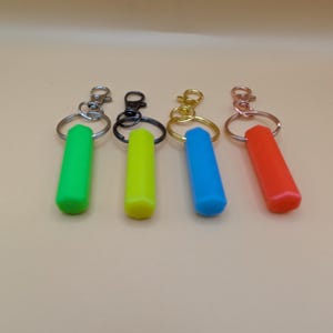 May include: Four keychains with colorful, cylindrical charms. The charms are green, yellow, blue, and red. Each keychain has a different metal clasp: silver, black, gold, and copper. The keychains are arranged on a light brown surface.