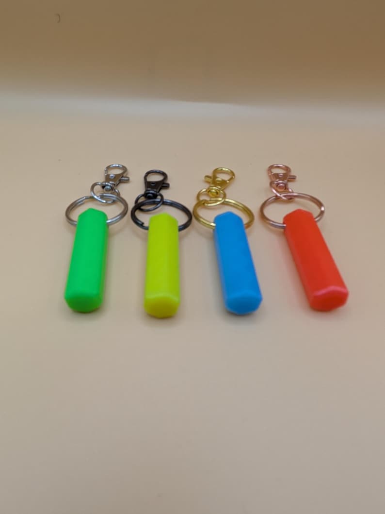 May include: Four keychains with colorful, cylindrical charms. The charms are green, yellow, blue, and red. Each keychain has a different metal clasp: silver, black, gold, and copper. The keychains are arranged on a light brown surface.