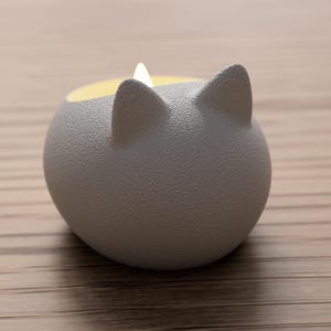May include: A gray, cat-shaped candle holder with a textured surface. The candle holder has two pointed ears and a yellow interior, with a small, lit candle inside. The holder is sitting on a light brown wooden surface.