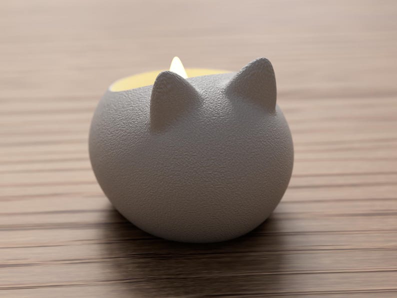 May include: A gray, cat-shaped candle holder with a textured surface. The candle holder has two pointed ears and a yellow interior, with a small, lit candle inside. The holder is sitting on a light brown wooden surface.