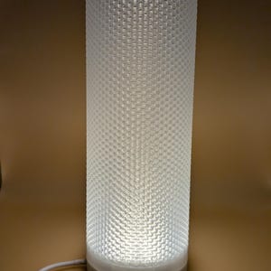 K&ouml;nnte beinhalten: Eine zylindrische wei&szlig;e Lampe mit einem strukturierten, wabenartigen Muster. Die Lampe wird von innen beleuchtet und wirft ein sanftes Licht. Der Sockel besteht aus einem hellen Material, und ein wei&szlig;es Kabel erstreckt sich von der Basis.