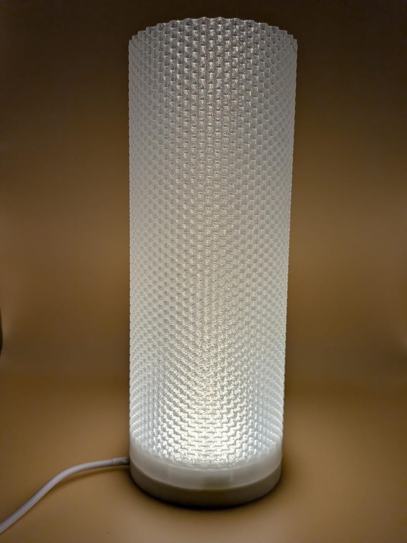 K&ouml;nnte beinhalten: Eine zylindrische wei&szlig;e Lampe mit einem strukturierten, wabenartigen Muster. Die Lampe wird von innen beleuchtet und wirft ein sanftes Licht. Der Sockel besteht aus einem hellen Material, und ein wei&szlig;es Kabel erstreckt sich von der Basis.