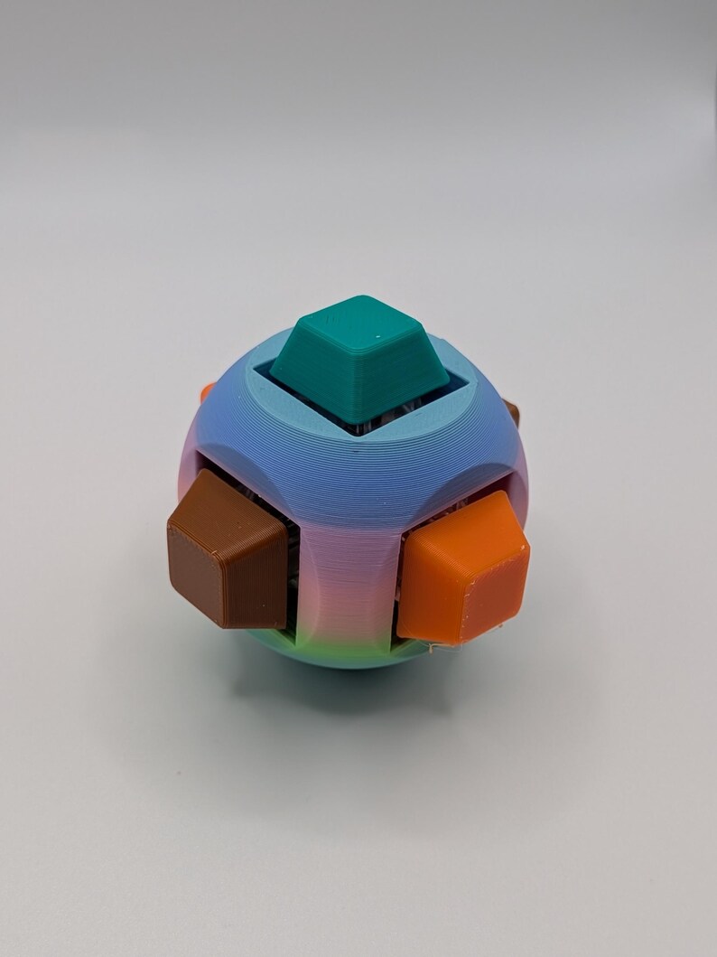 May include: A colorful 3D-printed puzzle toy. The spherical shape features a gradient of blue, pink, and green. It has six cube-shaped pieces in teal, brown, and orange. The toy is designed with a unique, geometric aesthetic.