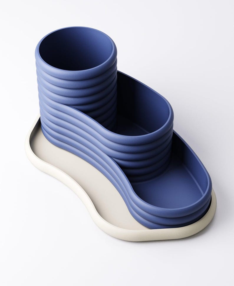 May include: A modern desk organizer in a minimalist design. The organizer is a deep blue color with a cylindrical pen holder and a wavy tray. The base is a light beige color. The design is made of a smooth, solid material.