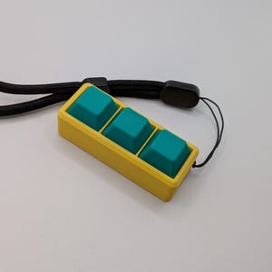 May include: A yellow rectangular device with three teal square buttons. The device is attached to a black lanyard. The buttons are slightly raised and textured. The background is a plain white surface.