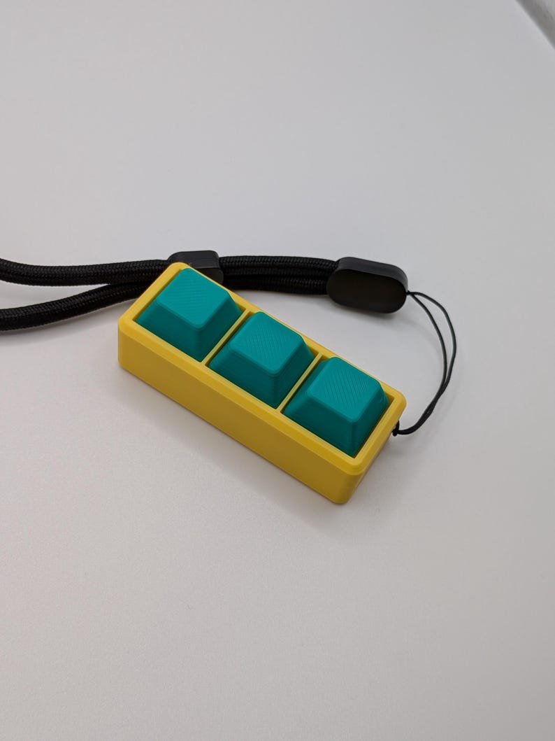 May include: A yellow rectangular device with three teal square buttons. The device is attached to a black lanyard. The buttons are slightly raised and textured. The background is a plain white surface.