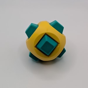 May include: A geometric puzzle toy with a yellow body and teal-colored square inserts. The toy has a symmetrical design with a central square and six protruding squares. The toy is designed to be manipulated and solved.