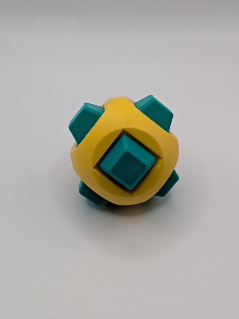 May include: A geometric puzzle toy with a yellow body and teal-colored square inserts. The toy has a symmetrical design with a central square and six protruding squares. The toy is designed to be manipulated and solved.
