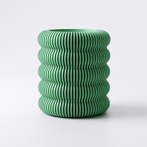 May include: A green, cylindrical vase with a unique design. The vase features a series of vertical, wavy lines that create a ribbed effect. The color is a soft, muted green, and the overall shape is rounded and organic. The vase is a decorative item.