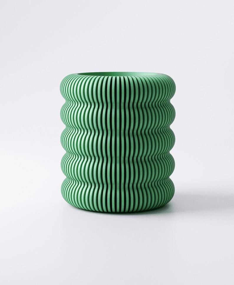 May include: A green, cylindrical vase with a unique design. The vase features a series of vertical, wavy lines that create a ribbed effect. The color is a soft, muted green, and the overall shape is rounded and organic. The vase is a decorative item.