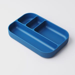May include: A blue plastic divided tray with four compartments. The tray has a ribbed exterior and rounded corners. The compartments are different sizes, suitable for organizing small items. The tray is a solid, vibrant blue color.