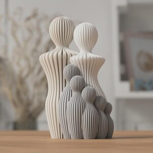 Puede incluir: Una escultura familiar de madera con un dise&ntilde;o minimalista. La escultura presenta dos figuras grandes y cuatro figuras m&aacute;s peque&ntilde;as, todas con un patr&oacute;n de rayas. Las figuras son en tonos de blanco y gris, y est&aacute;n dispuestas sobre una superficie de madera.