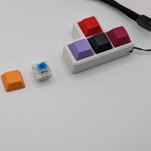 May include: A collection of colorful mechanical keyboard keycaps in red, black, purple, and magenta, arranged in a white holder. An orange keycap and a clear switch are also visible. A black wrist strap is in the background.
